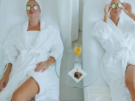 Two women in white robes and towels on their heads lay on chairs with cucumbers on their eyes
