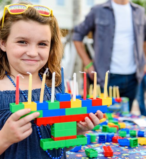 young girl with sunglasses in her hair holds up a menorah made out of multi colored blocks