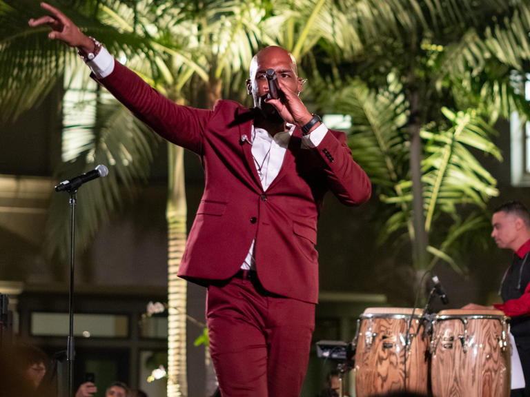 man in red suit holds microphone to mouth and points hand in air